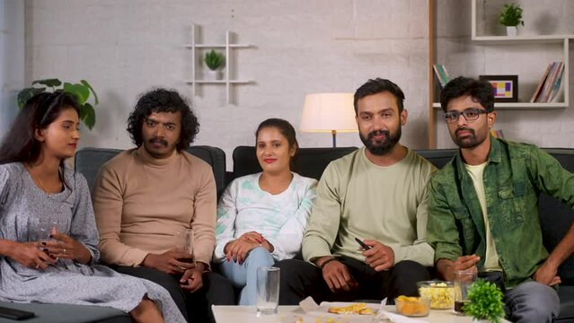 Group of relaxed friends watching movie on tv or television at home - concept of refreshments, weekend gathering or reunion and entertainment.