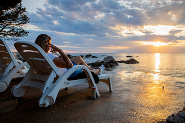 A woman is sitting on a deckchair on the seashore, having fun with her phone and enjoying the sunset