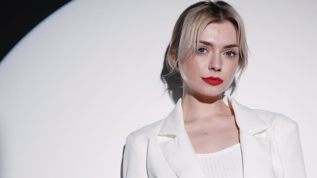 Young beautiful confident blond woman wearing trendy white suit and jacket. Fashion model posing in studio. Fashionable female posing near wall in a circle light. Cheerful and happy. With red lips