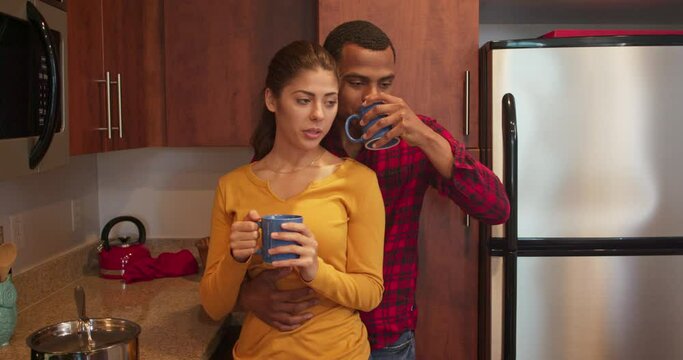 Millennial couple drinking morning coffee and playfully kissing in the kitchen. African American man drinking tea and holding Caucasian woman. 4k slow motion handheld