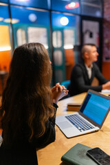 Brunette woman with loose hair sits crossing arms by modern laptop in creative office. Female thinks about business solutions plan