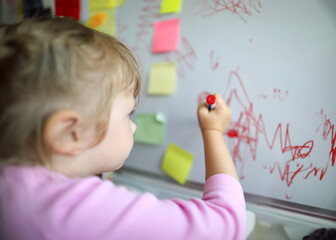 Focus on tender small hand of little girl holding big red marker and drawing cute childish paintings on glass board in modern classroom. Creative childhood concept