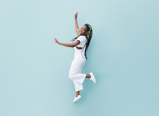 Happy woman in white casual clothes jumping against a blue wall. Young female with braids jumps outdoors. - obrazy, fototapety, plakaty