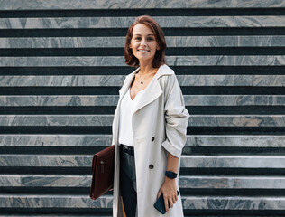 Cheerful middle-aged woman in stylish clothes looking at the camera while standing outdoors. Female...