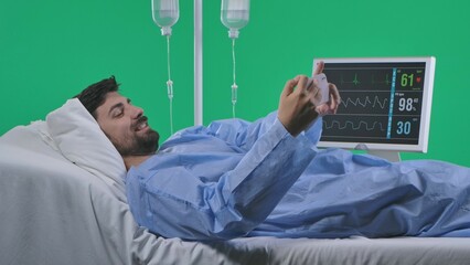 Adult man patient laying in bed with drip and monitor, holding smartphone and taking selfie, smiling face expression. Isolated on chroma key green screen.