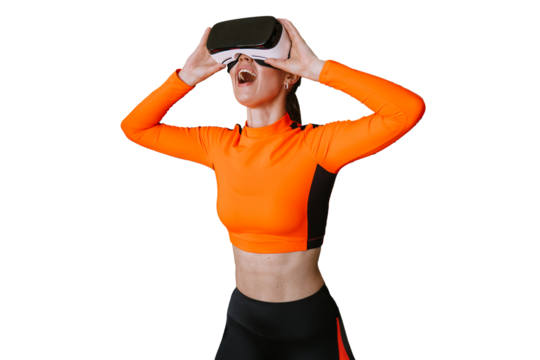 Excited young woman in orange sportswear using vr googles at gym surprised by technologies standing at gym with opened mouth against shelves with fitness equipment.