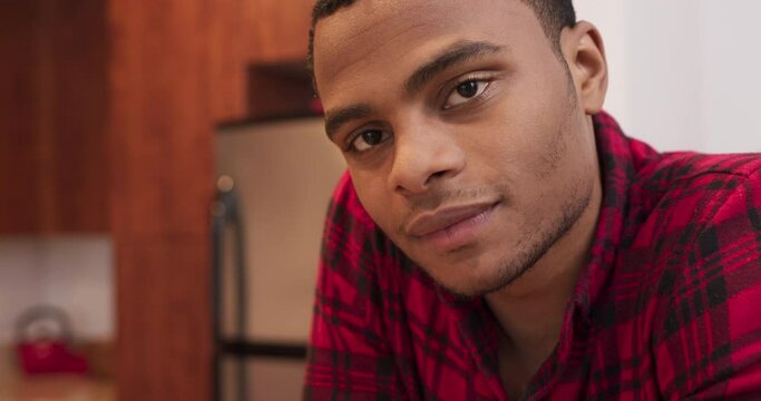 Close up portrait of attractive young black man looking at camera and smiling. African American Millennial guy at home in red plaid short. 4k slow motion handheld