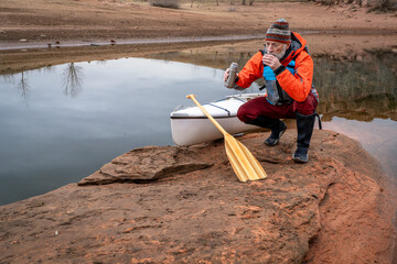 hot tea break - senior male paddler with a decked expedition canoe with a wooden paddle on a rocky shore of Horsetooth Reservoir near Fort Collins, Colorado, low water level fall or winter scenery - obrazy, fototapety, plakaty