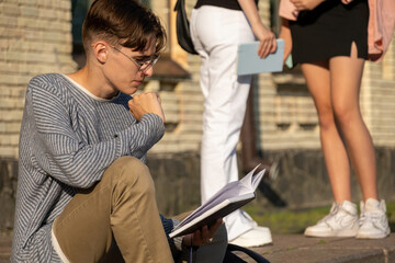 Young man enthusiastically reads book while sitting near university building. Guy with glasses...
