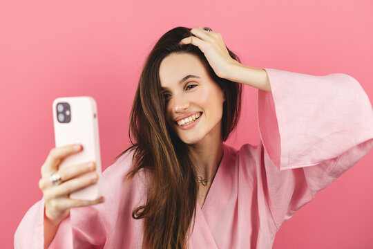 Brunette girl stares at the phone and fixes her hair. Female uses phone like mirror. Young girl make photo from hands with phone on pink background.