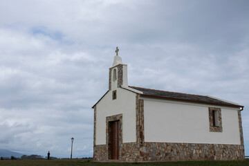 A small white chapel with a stone base under a cloudy sky, next to a grass field and a lamppost