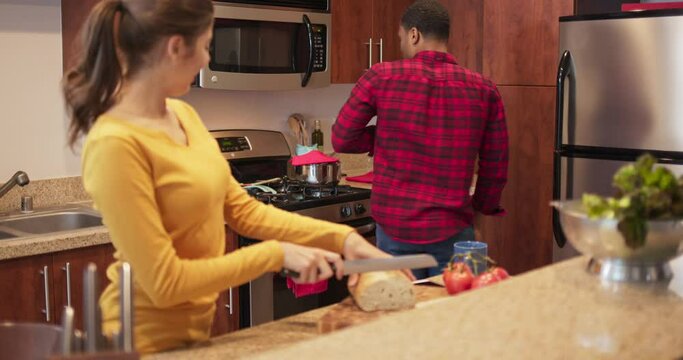 Caucasian woman laughing and  cutting bread while her African American boyfriend cooks marinara in the background then dances over to her. Millennial newlyweds making dinner. 4k slow motion handheld