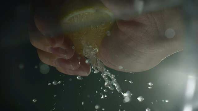 Hand of cook squeezing a lemon juice in slow motion. Macro of splash a citrus limonum juice against black background. Cooking food and drink concept