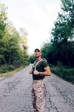 Army man in uniform with backpack and cap standing on road against trees in forest