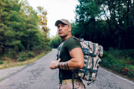 Army man in uniform with backpack and cap standing on road against trees in forest