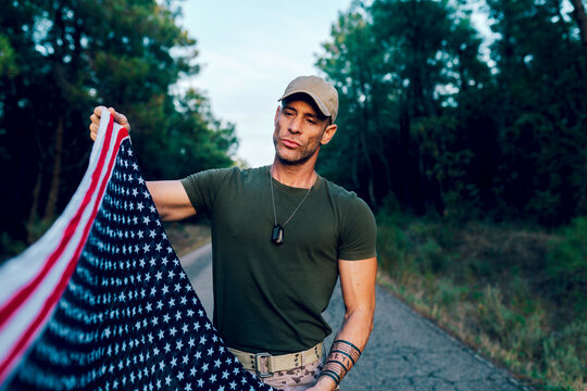 Military soldier holding and looking at national flag with pride while standing on road against forest