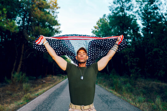 Military soldier with closed eyes waving national flag with pride while standing on road against forest