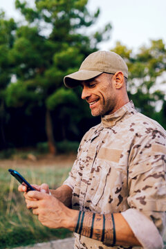 Side view of Happy mature army soldier with smartphone against forest