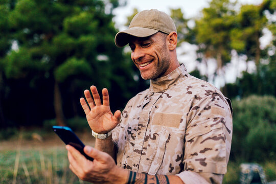 Smiling mature commando waving at mobile phone while standing in forest