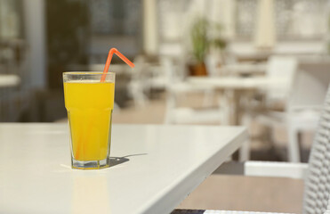 Glass of orange juice with plastic straw on white table in restaurant outdoor lounge zone in sunny weather