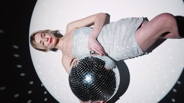 Vertical video. Young beautiful smiling blond female in trendy evening silver dress. Sexy woman posing near white wall in a circle of light. Fashionable model with bright makeup. Holds disco ball