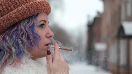 A woman with bright colored hair, wearing a white fur coat, smokes a cigarette outdoors in winter....