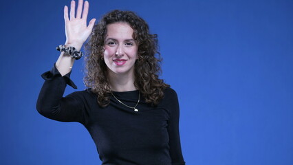 Happy woman greeting to camera with hand gesture standing on blue background. 20s Person saying...