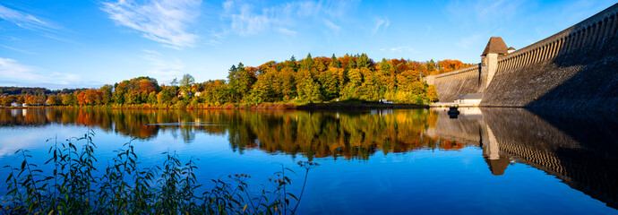 Panoramic view of Möhne dam and  pond below the famous dam brick wall on sunny autumn day with colorful foliage and reflection. Idyllic recreation area at village of “Günne“ perfect for hiking. - Powered by Adobe