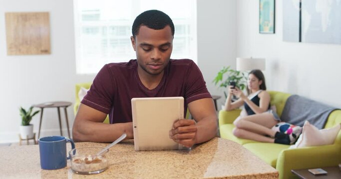 Black millennial man eating breakfast at the kitchen counter while checking his tablet computer with his girlfriend on the couch using her smartphone in the background. 4k slow motion handheld