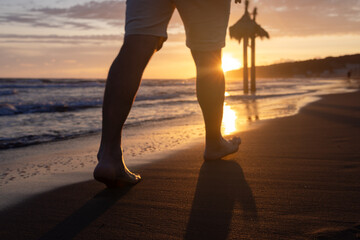 The legs of a man walking barefoot on a sandy beach. A sunny path