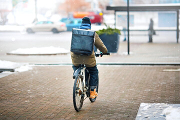 Food delivery man riding bicycle on snowy city street with large black backpack. Delivery boy on...
