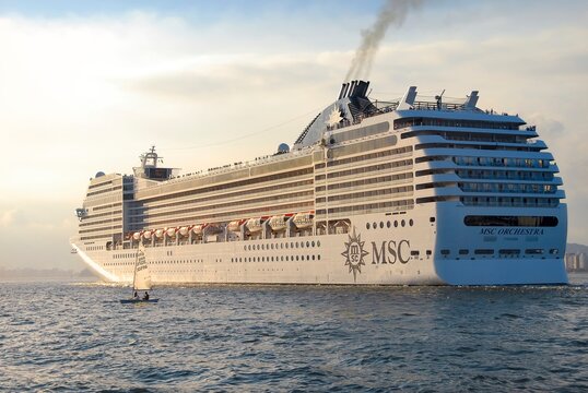 Cruise ship MSC Orchestra sailing during sunset. Sailboat next to the ship. City buildings in the background. Santos city, Brazil. 