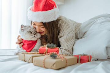 Happy asian woman wearing Santa hat playing grey cat wearing red scarf. Friendship with pets in...