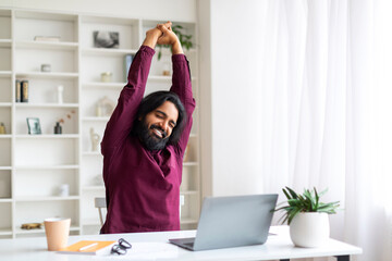 Relaxed Indian freelancer man taking break and stretching after working on laptop