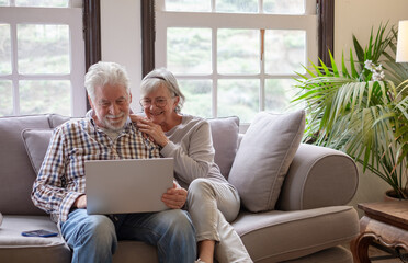 Happy 70s white haired senior couple sitting on sofa in living room using laptop surfing the net. Concept of older generation users and wireless technology - obrazy, fototapety, plakaty