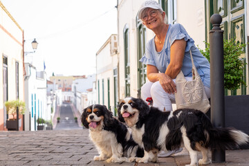 Smiling elderly woman with hat sitting on the street with her two cavalier king charles spaniel...