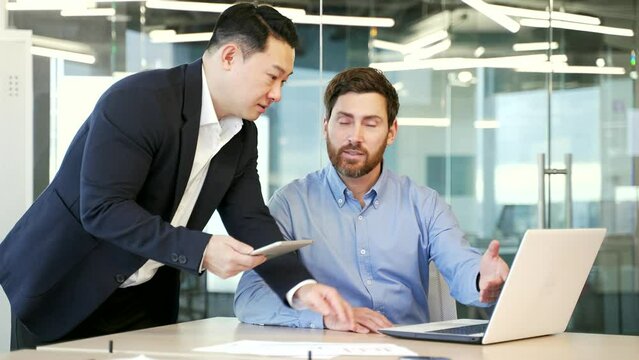 Two diverse colleagues discussing new project, businessmen using digital tablet and laptop compter together in office. professional man corporate employees in conversation working on business project