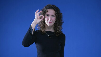 Person giving thumbs up to camera while standing on blue background. Woman showing OK sign with...