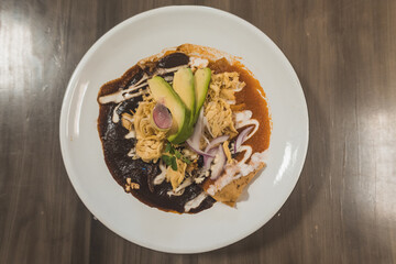Red enchiladas combined with mole enchiladas, accompanied with chicken, cream, cheese, avocado, on a white plate, on a wooden table.