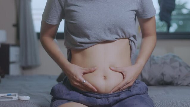 Close-up shot of her hand touching and shaking her fat belly and cellulite of an obese Asian womanin bed. The stretched skin on her abdomen emphasizes her weight gain and concerns about her health. 