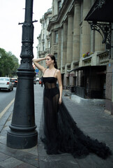 A 30-year-old girl in a black transparent revealing dress on a city street. Style and elegance, femme fatale, photo shoot, advertising, girl in a black haute couture dress, close-up