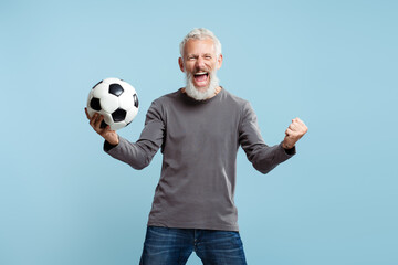 Bearded attractive mature man holding soccer ball cheering, standing isolated on blue background