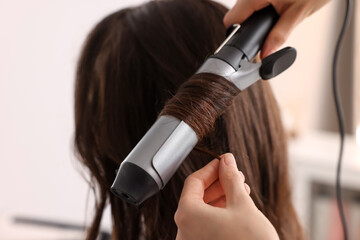 Hairdresser working with client using curling hair iron in salon, closeup