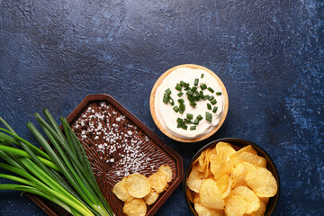 Bowl of tasty sour cream with sliced green onion and potato chips on blue background