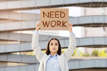A determined young woman in business attire holding a sign that reads NEED WORK