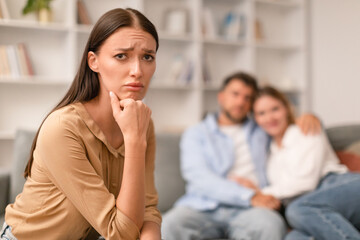 Sad Woman Sitting While Her Ex Boyfriend Hugging Girlfriend Indoors