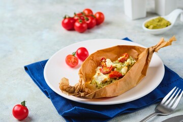 Fish baked with pesto sauce, cherry tomatoes and feta in portioned parchment forms on a white plate on a gray concrete background. Fish recipes.