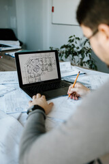 An arhitect working in his office, on his laptop and using a pen, designing a new project.