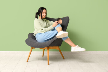 Fototapeta na wymiar Beautiful young woman with mobile phone sitting on comfortable armchair near green wall