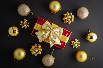 New Year concept. Top view photo of stylish red giftbox with golden ribbon bow and Christmas decorations balls on isolated black background with copyspace.
